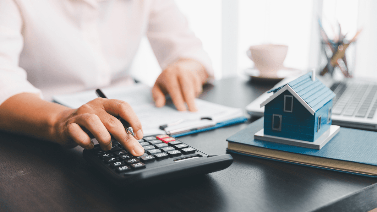 Person using a calculator to compare mortgage refinance options beside a model home and financial documents.