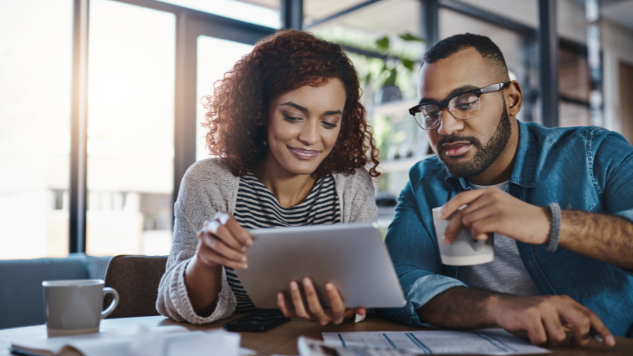 Smiling couple sitting at a table, reviewing something on a tablet together with coffee and documents nearby.