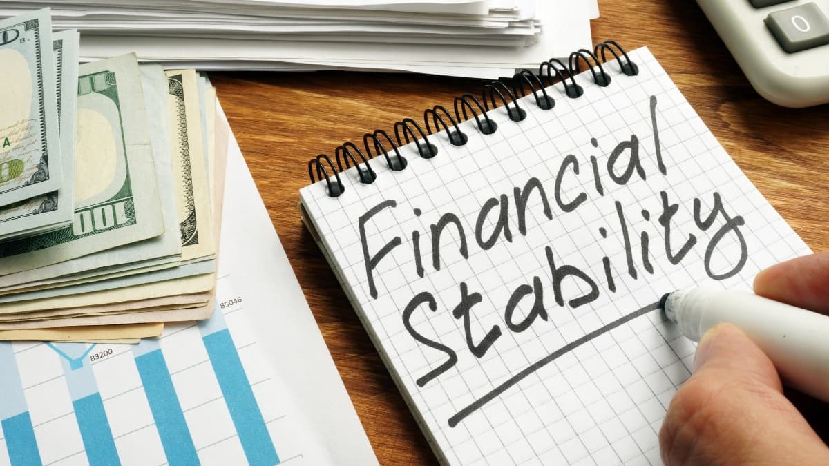 A person writing “Financial Stability” on a notepad beside cash, financial charts, and a calculator, symbolizing smart money habits and personal finance tips for saving and budgeting.